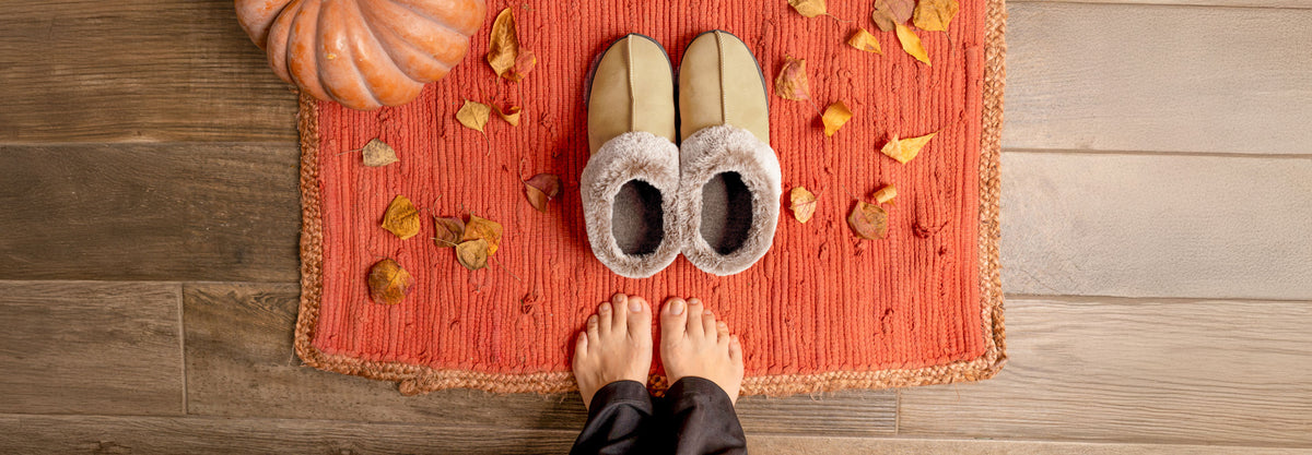 Pair of slippers on a orange rug with autumn leaves and a pumpkin on a wooden floor.
