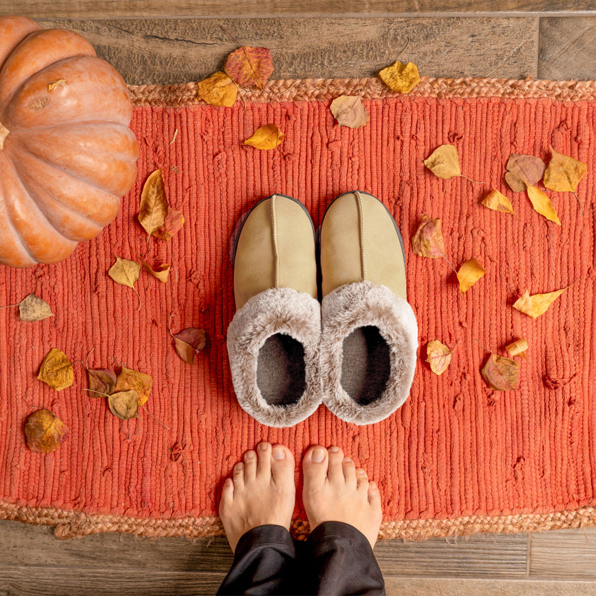 Pair of slippers on a orange rug with autumn leaves and a pumpkin on a wooden floor.